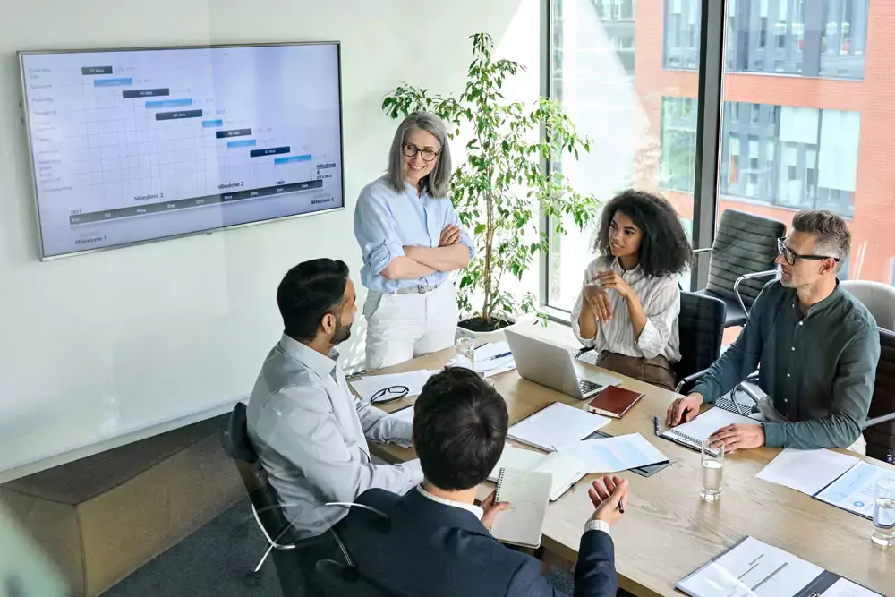 Senior female ceo and multicultural business people discussing company presentation at boardroom table with a gantt chart on a screen.